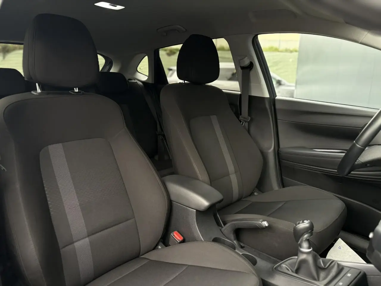 Vue intérieure côté droit de la Hyundai i20 2025, sièges tissu noirs avec accoudoir central et levier de vitesses manuel.