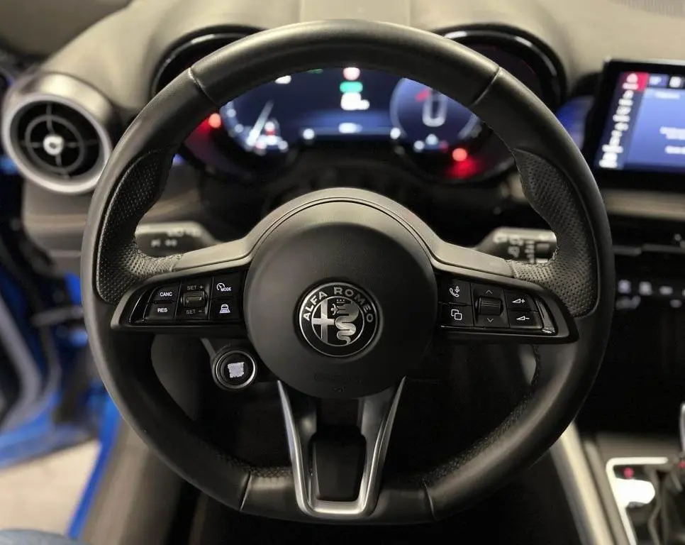 Volant sport en cuir noir avec logo Alfa Romeo au centre, tableau de bord digital et partie de la portière bleu visible.