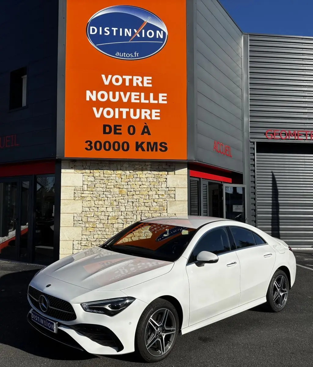 Mercedes CLA 250 e AMG Line blanc vue 3/4 avant droit, stationnée devant un bâtiment avec panneau orange Distinxion.