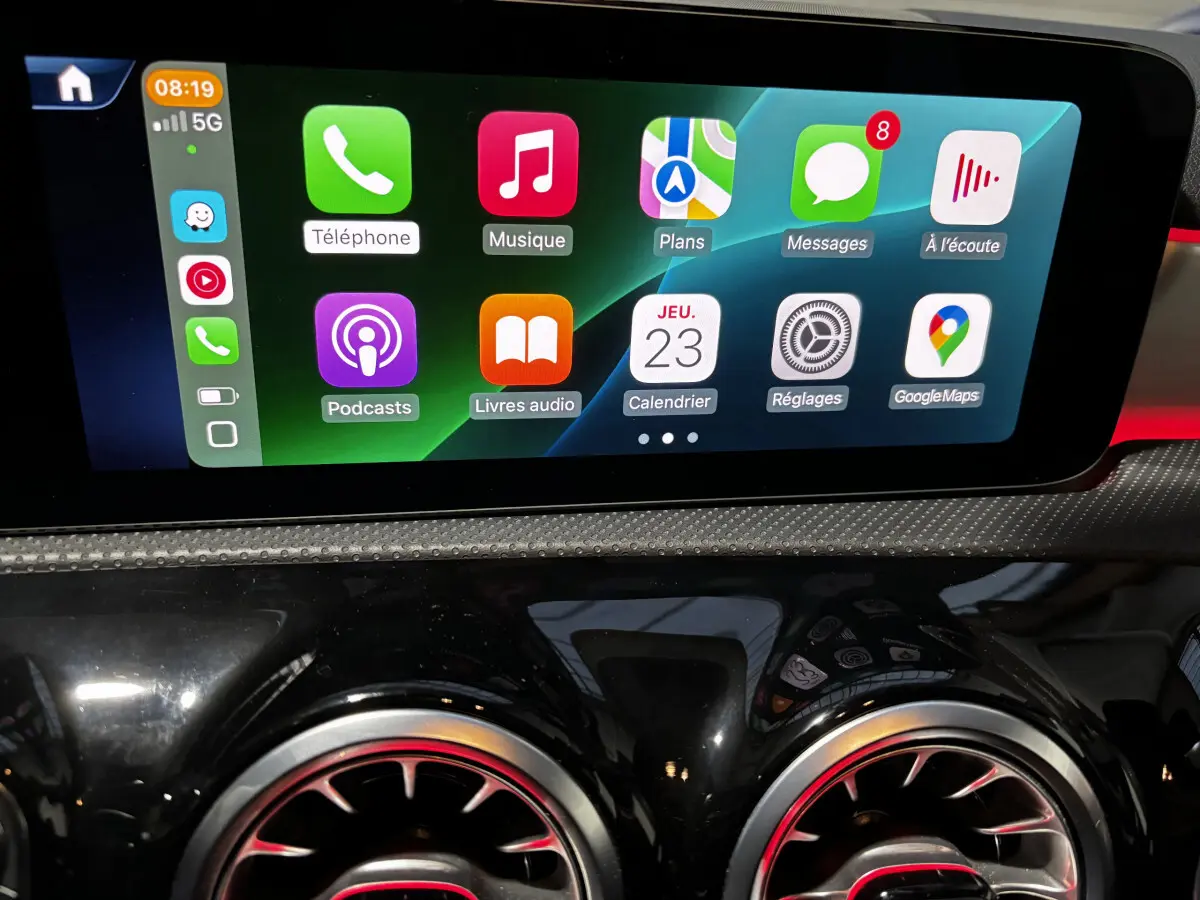 Écran tactile central affichant des applications, avec sorties d'air rondes noires et touches rouges en reflet sur tableau de bord noir brillant.