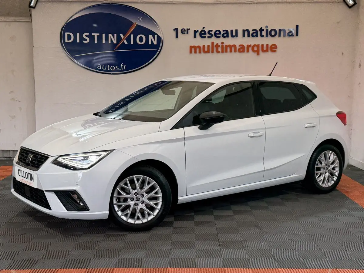 Vue 3/4 avant droite d'une SEAT Ibiza blanche 2024 avec jantes alliage et phares LED allumés en intérieur showroom.