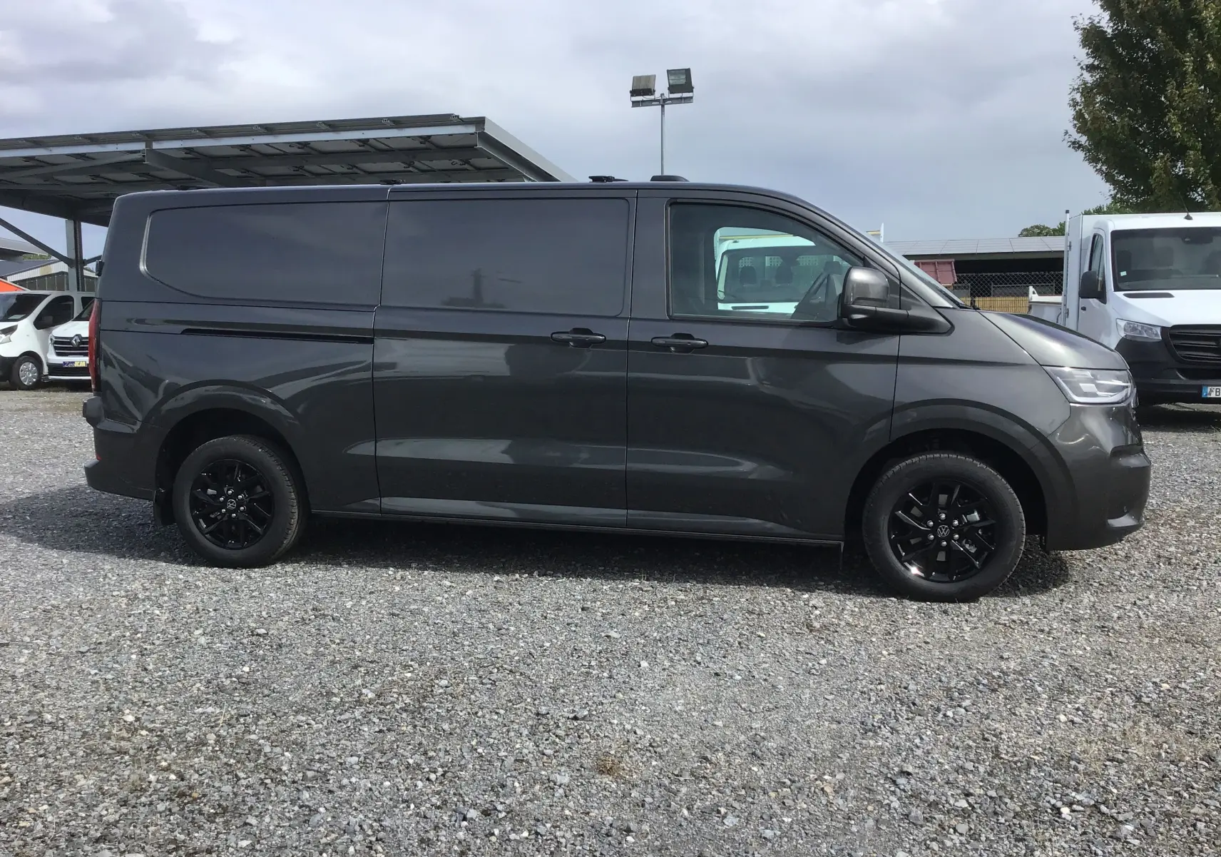 Profil côté gauche d'un Volkswagen Transporter T7 gris anthracite avec jantes noires, stationné sur un sol gravillonné.