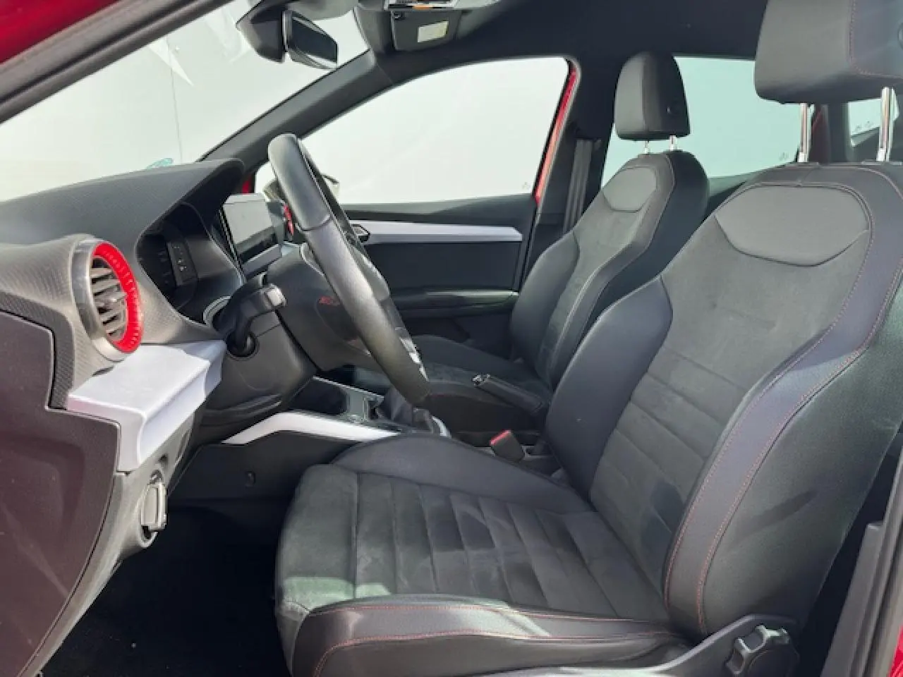 Vue intérieure côté conducteur du SEAT Arona 2024, sièges en suédine noire avec surpiqûres rouges et volant sport cuir.