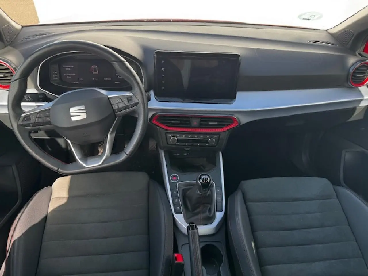 Vue intérieure frontale du SEAT Arona 2024 avec volant cuir, console centrale manuelle et touches rouges décoratives.