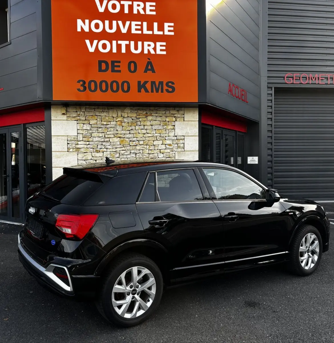 Audi Q2 noir vue 3/4 arrière droit, stationné devant un bâtiment avec panneau orange et pierre apparente.