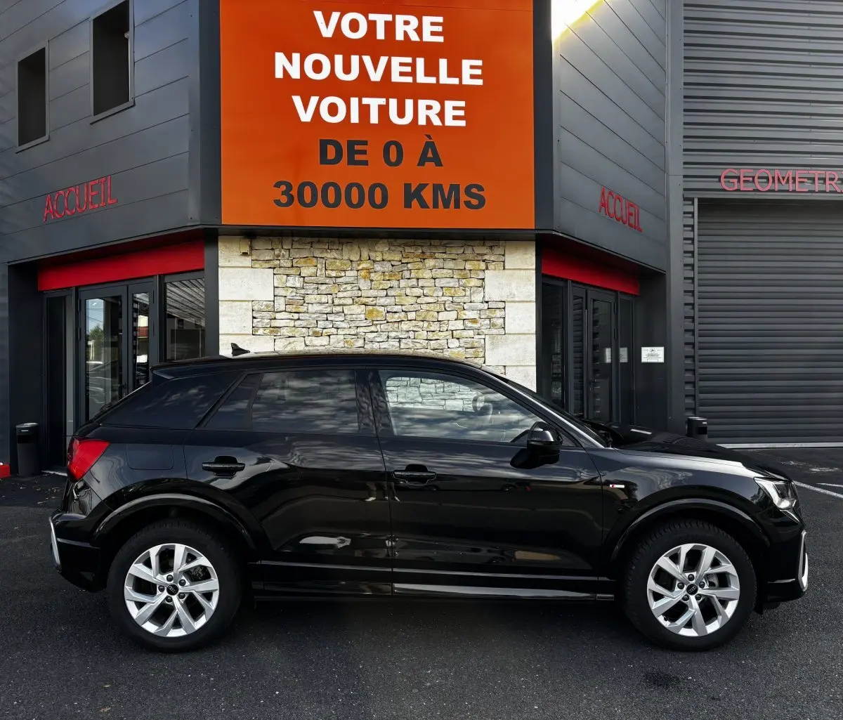 Audi Q2 35 TFSI S Line noir vue de profil côté gauche, stationné devant un bâtiment avec panneau orange.
