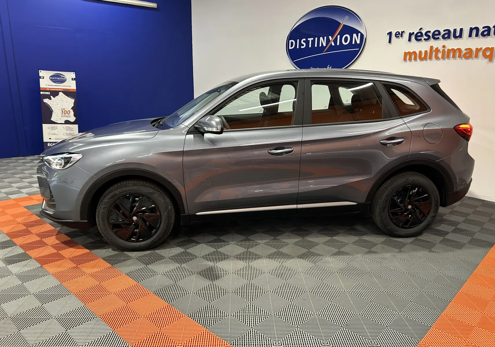 Vue de profil côté gauche du SUV MG ZS Hybrid+ 2025 en gris Hampstead avec jantes noires, dans un showroom.