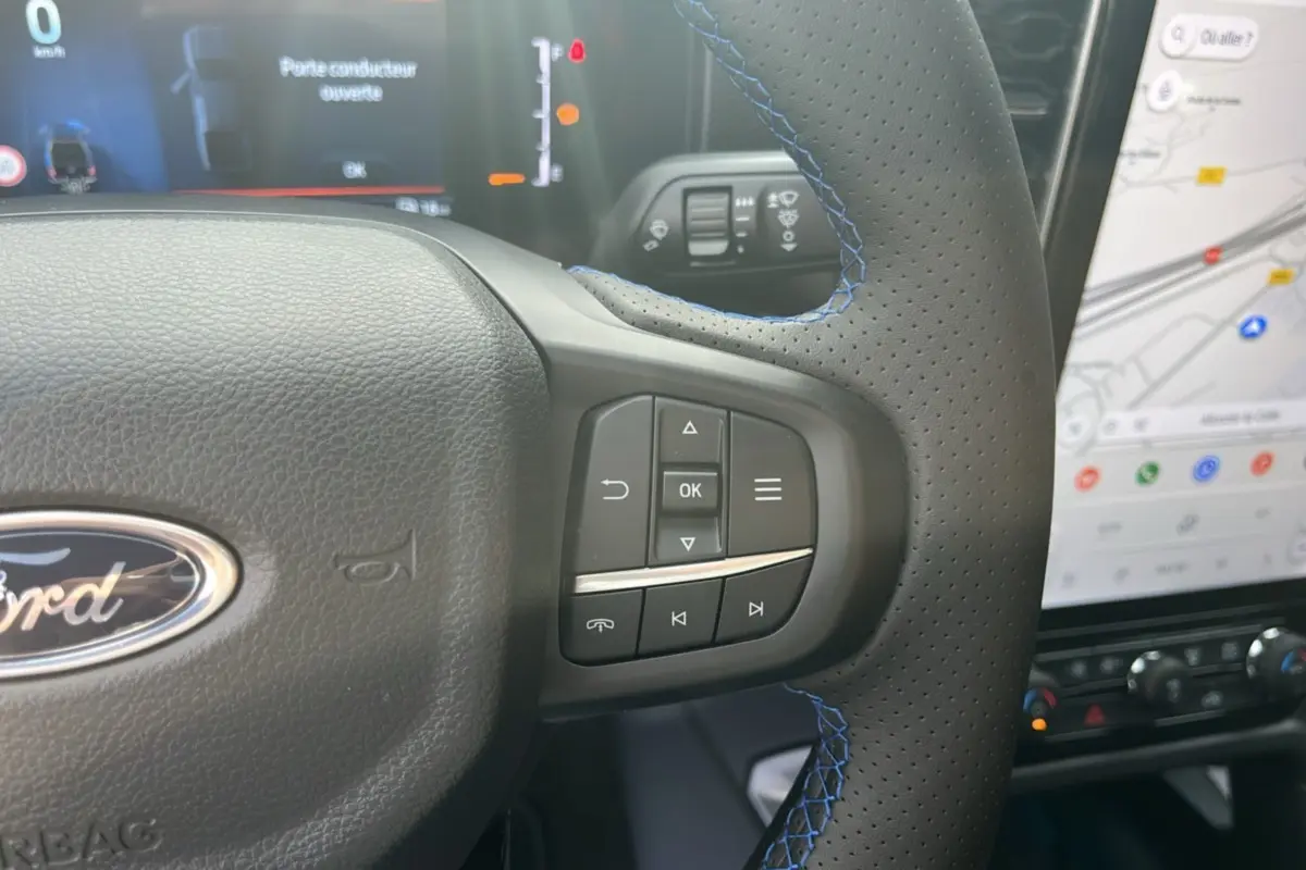 Gros plan sur le volant cuir perforé avec surpiqûres bleues du Ford Ranger 2025, écran tactile et tableau de bord en arrière-plan.
