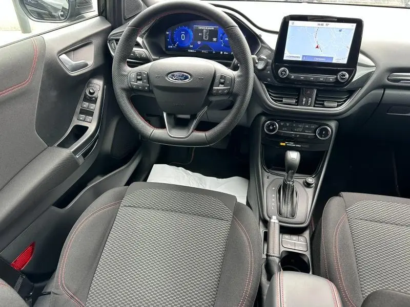 Vue intérieure avant du Ford Puma 2024, tableau de bord avec écran tactile, volant multifonction et sièges tissu noir avec surpiqûres rouges.