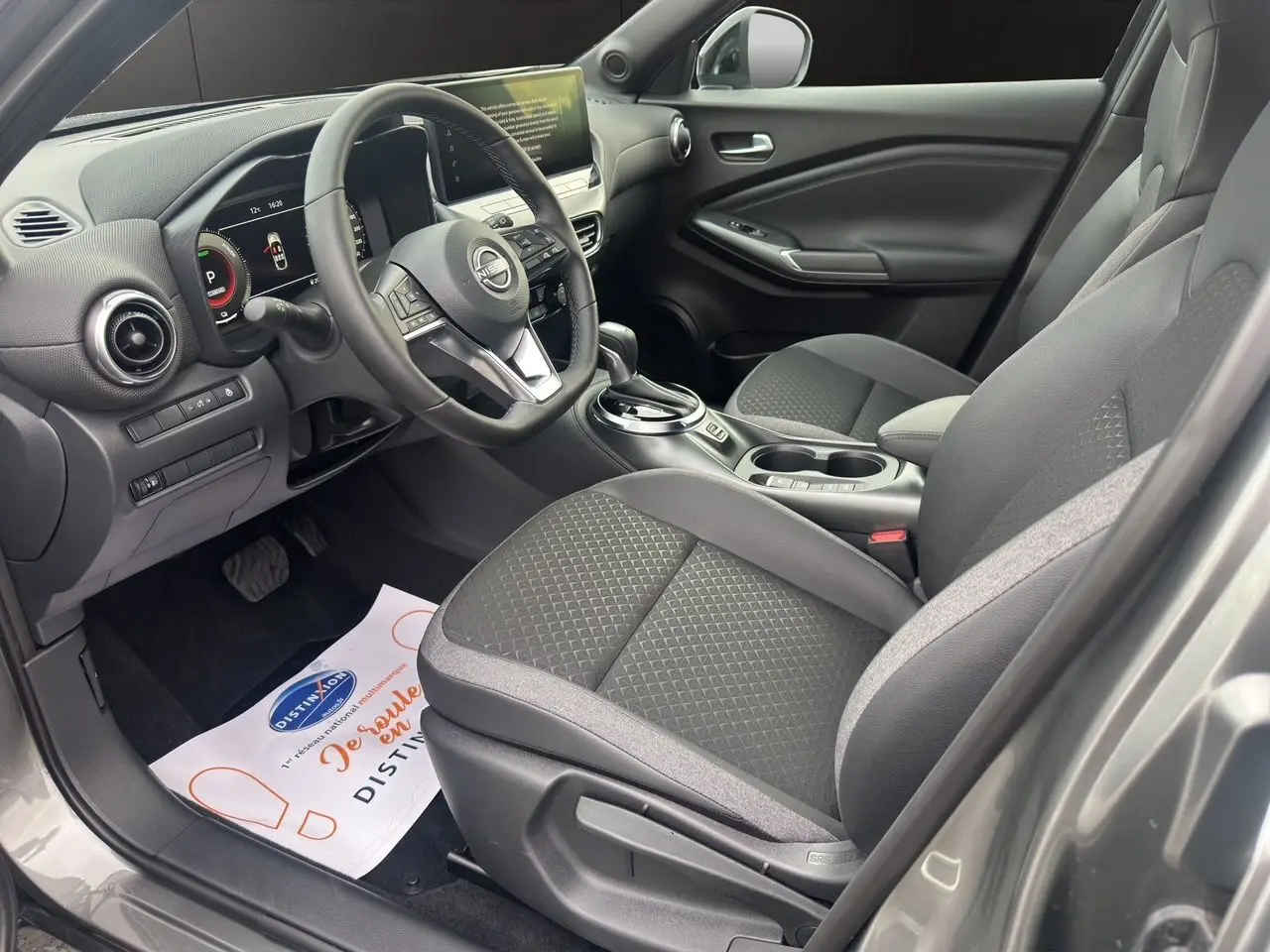 Vue intérieure côté conducteur du Nissan Juke Full Hybrid 2025 gris squale, avec sièges tissu noir et tableau de bord digital.