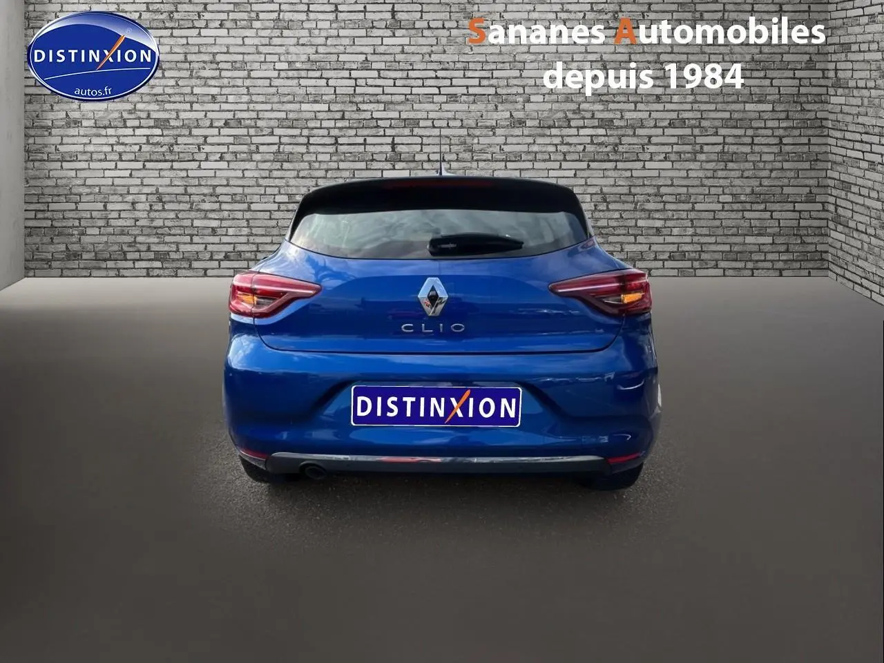 Vue arrière d'une Renault Clio 2021 bleu Iron, avec feux allumés et logo distinctif sur un fond de mur en briques.