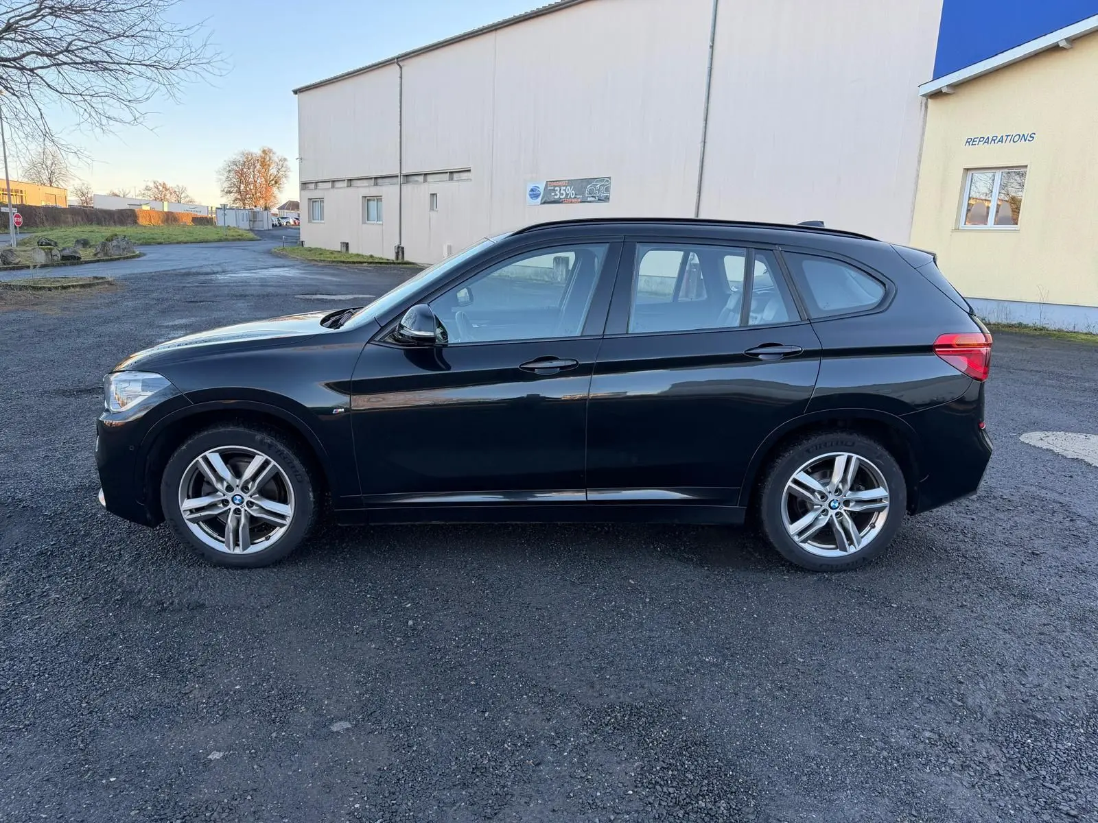 BMW X1 18D M Sport noir métal vue de profil côté gauche sur un sol goudronné devant un bâtiment clair.