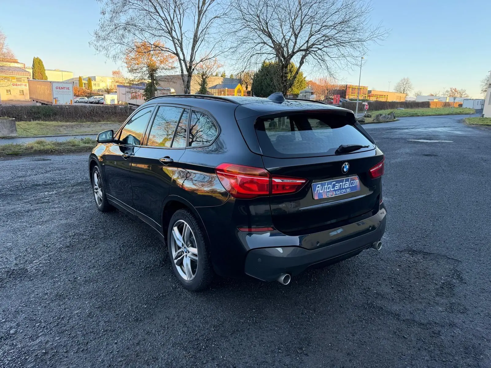 BMW X1 noir métal vue 3/4 arrière droit sur parking, avec feux arrière allumés et jantes alliage 18 pouces.