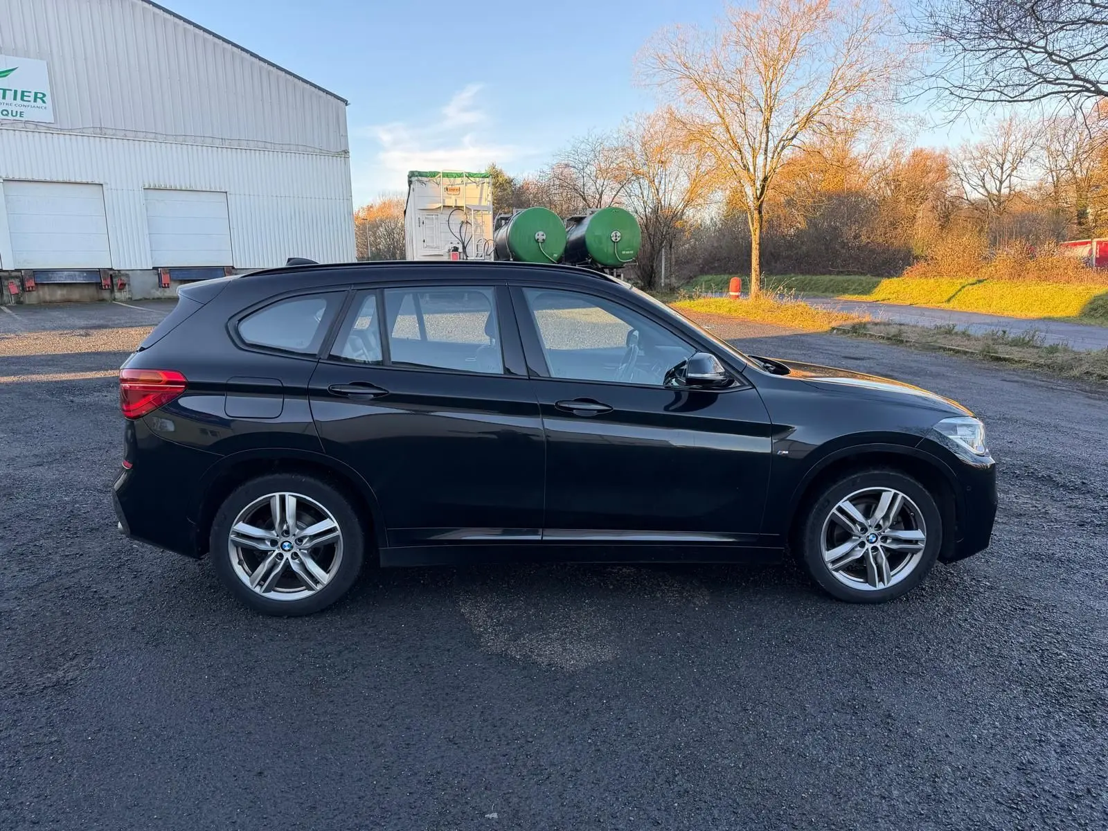 BMW X1 18D M Sport noir métal vue de profil côté gauche sur un sol goudronné en extérieur au coucher du soleil.