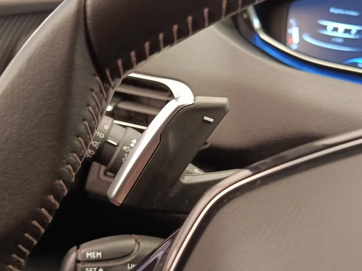 Gros plan sur la palette de changement de vitesse derrière le volant cuir d'une Peugeot 5008 gris foncé, tableau de bord partiellement visible.