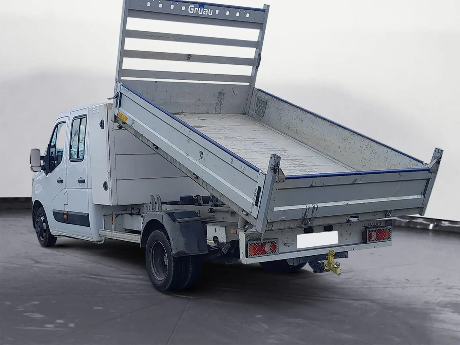 Renault Master blanc en double cabine vu 3/4 arrière avec benne basculante relevée et attelage visible.