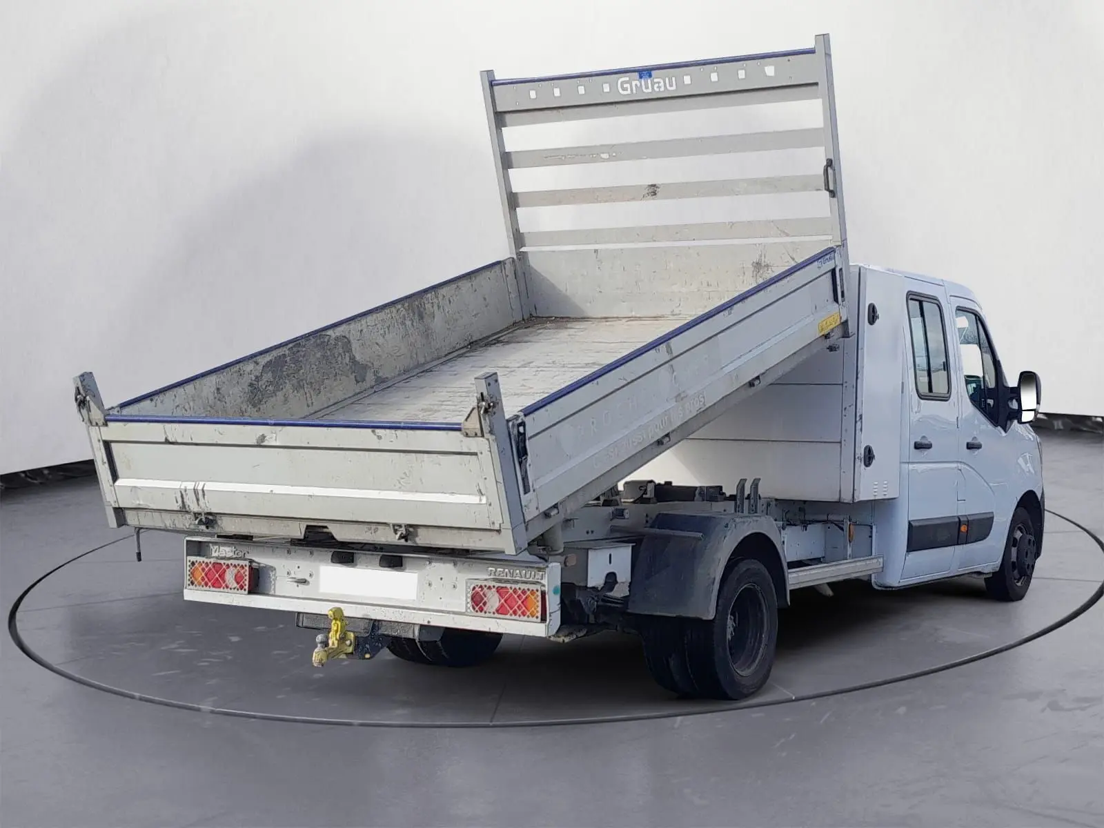 Renault Master blanc double cabine vu 3/4 arrière avec benne basculante relevée et attelage visible.