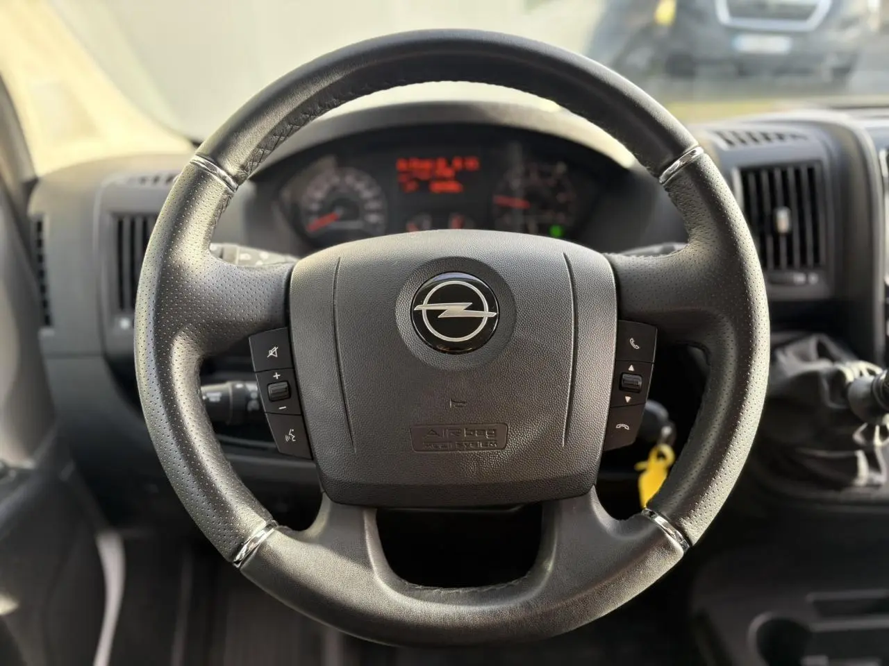 Vue rapprochée du volant noir perforé de l’Opel Movano Fourgon 2024 avec commandes intégrées et tableau de bord flou en arrière-plan.