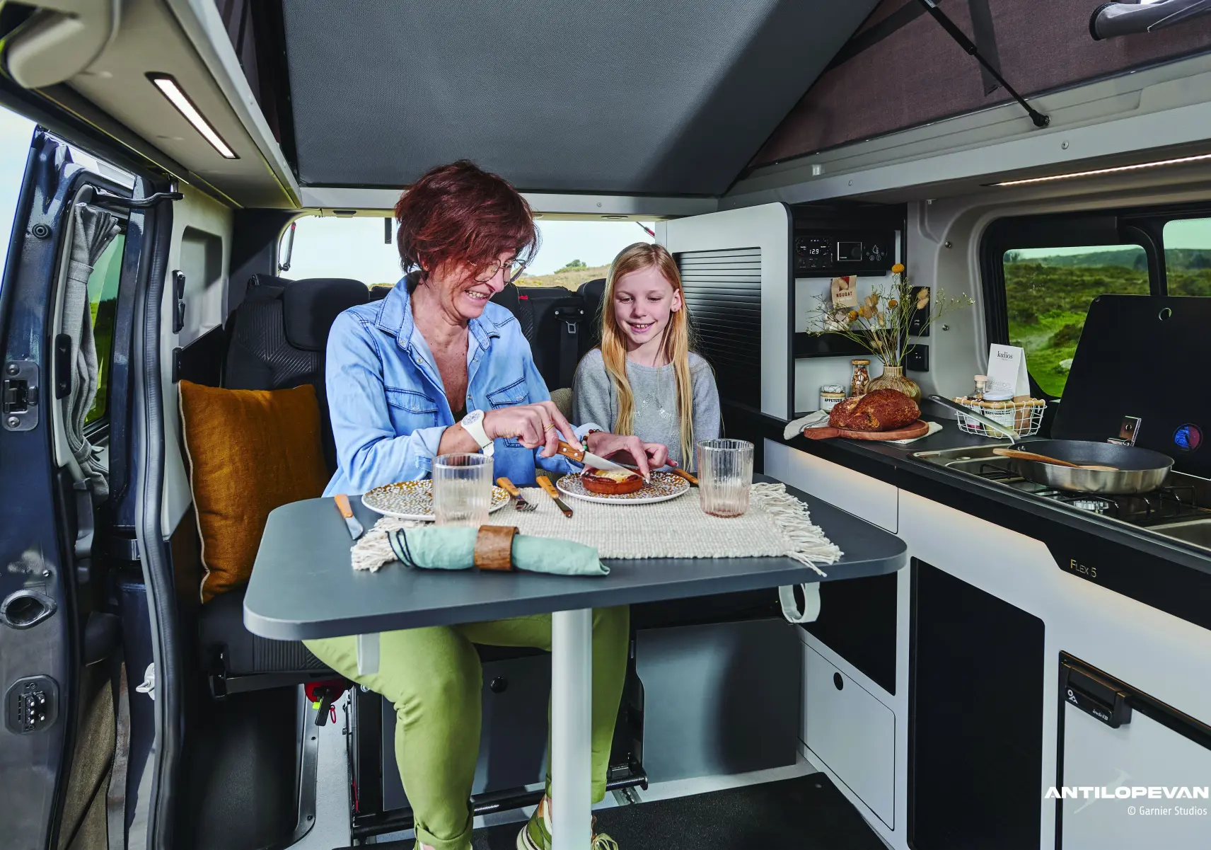 Intérieur du Renault Trafic Fourgon blanc glacier, vue latérale droite sur la cuisine aménagée et table avec deux personnes assises.