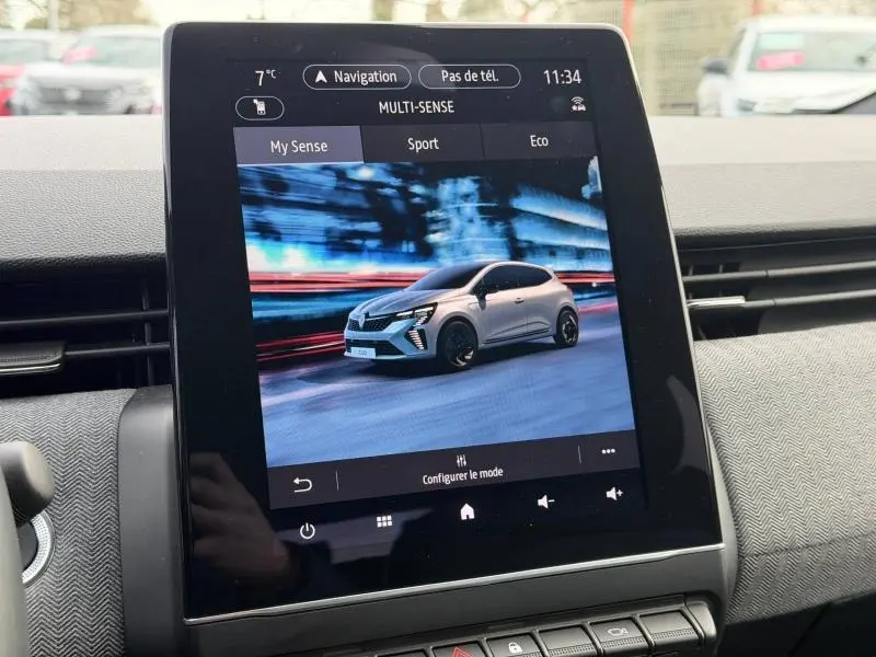 Écran tactile intérieur montrant une Renault Clio blanche en 3/4 avant sur fond urbain flouté.