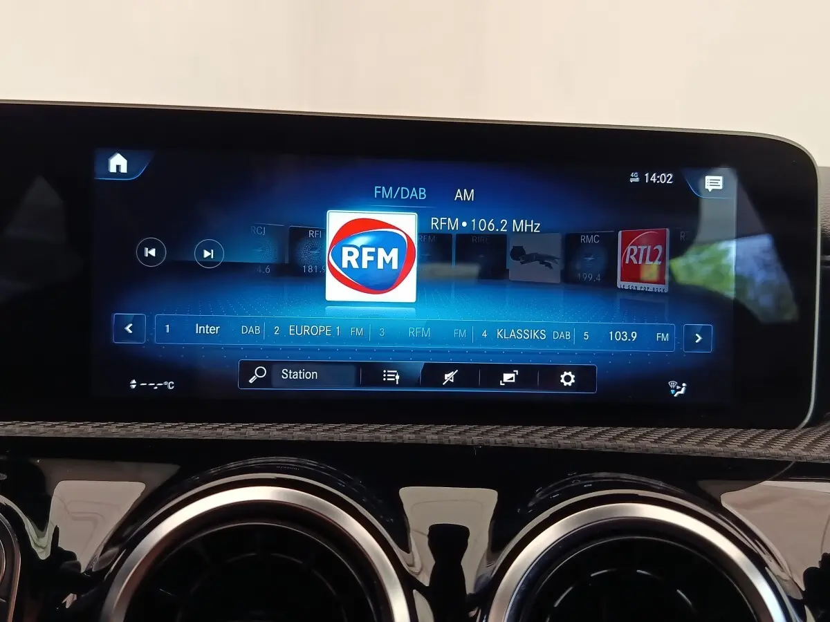 Écran tactile central affichant la radio RFM dans l'habitacle d'une Mercedes Classe A gris clair, vue de face.