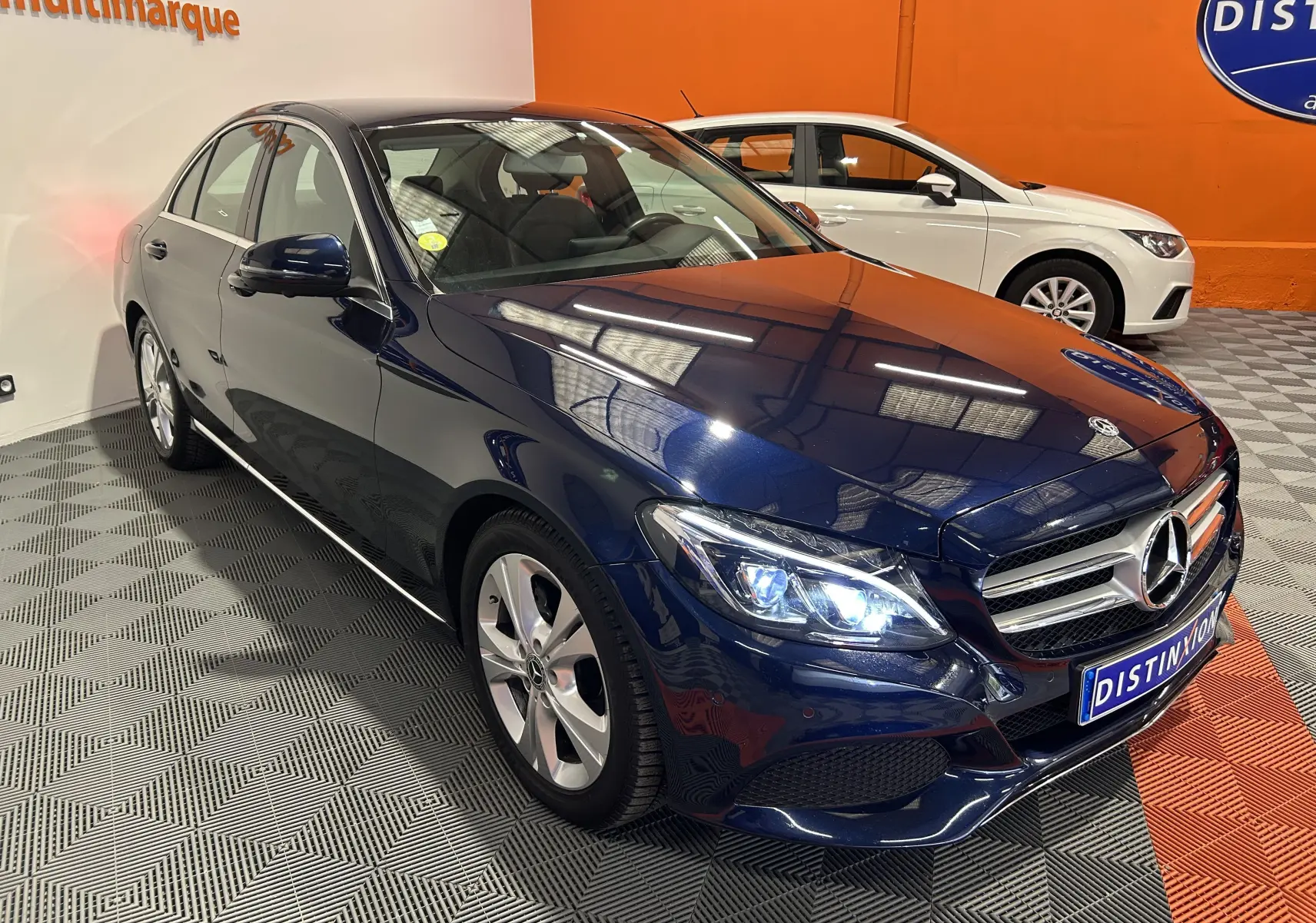 Mercedes Classe C 200d bleu vue 3/4 avant droit, phares allumés, dans un showroom avec sol damier gris et orange.