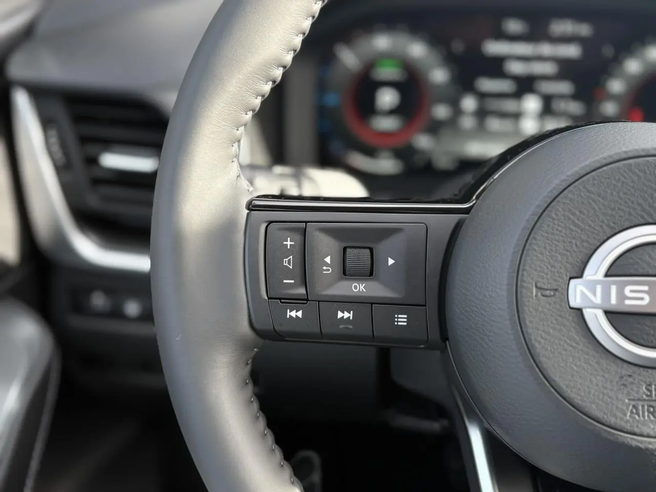 Gros plan sur les commandes gauche du volant cuir gris du Nissan Qashqai blanc nacré 2025 avec tableau de bord numérique flou.