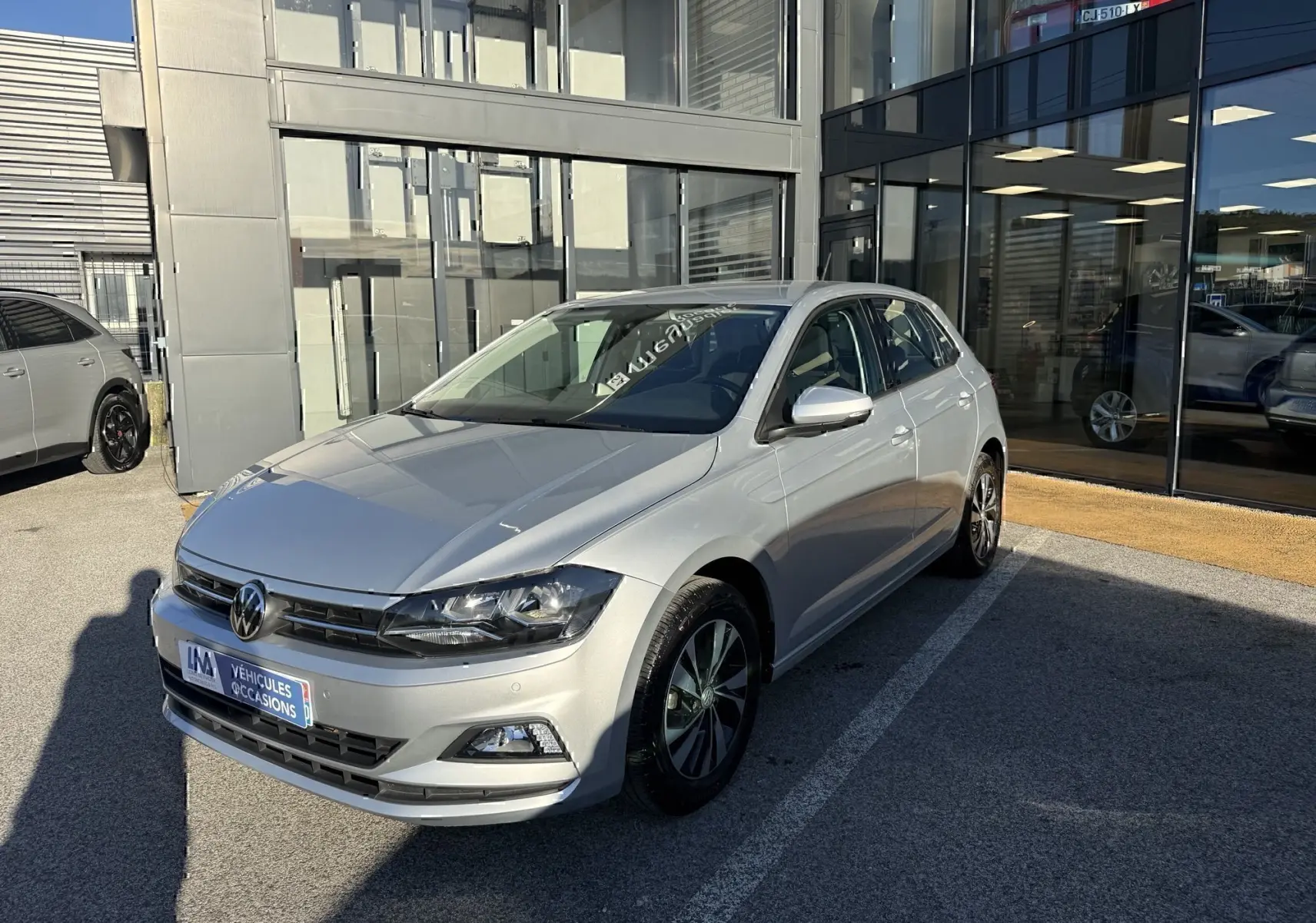 Volkswagen Polo gris métal vue 3/4 avant droit, stationnée devant un bâtiment vitré en plein jour.