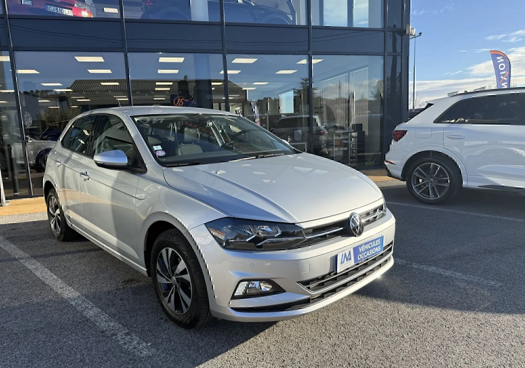 Volkswagen Polo gris métal vue 3/4 avant droit, avec jantes alliage et calandre noire brillante, stationnée devant un showroom.