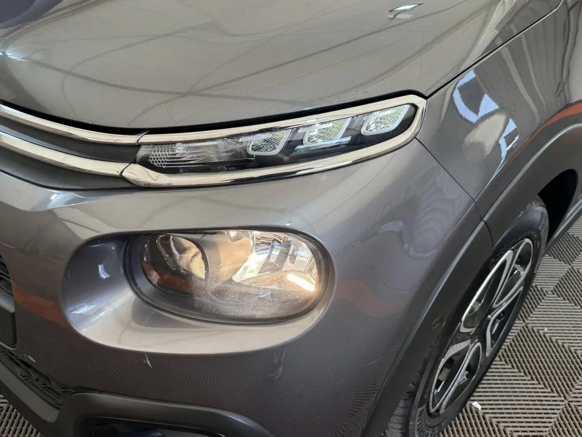 Gros plan sur le phare avant droit et la calandre chromée d'une Citroën C3 gris foncé, avec jante visible.