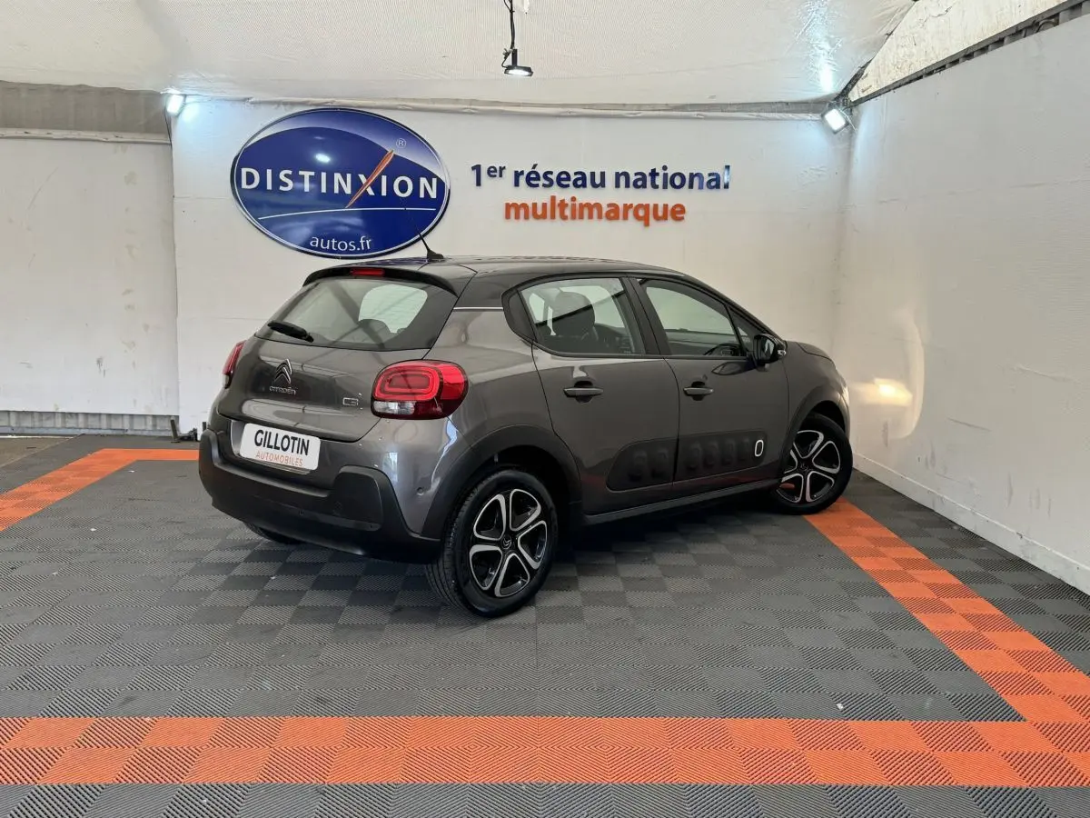 Vue 3/4 arrière droite d'une Citroën C3 gris foncé avec protections latérales noires et jantes bicolores dans un showroom.