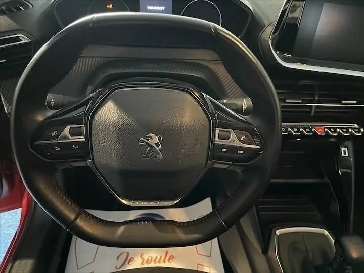 Vue rapprochée du volant multifonction noir de la Peugeot 208 rouge Elixir Metal, avec tableau de bord et console centrale visibles.
