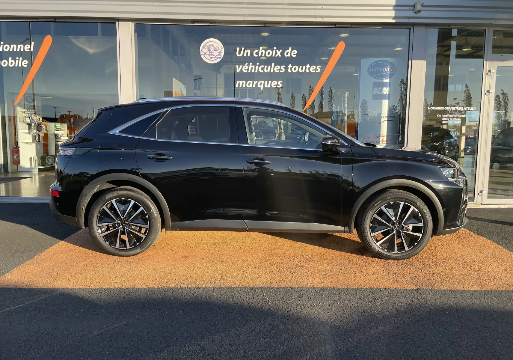 Profil latéral droit d'un DS7 noir Perla Nera 2024 avec jantes bi-ton et vitres teintées devant un showroom.