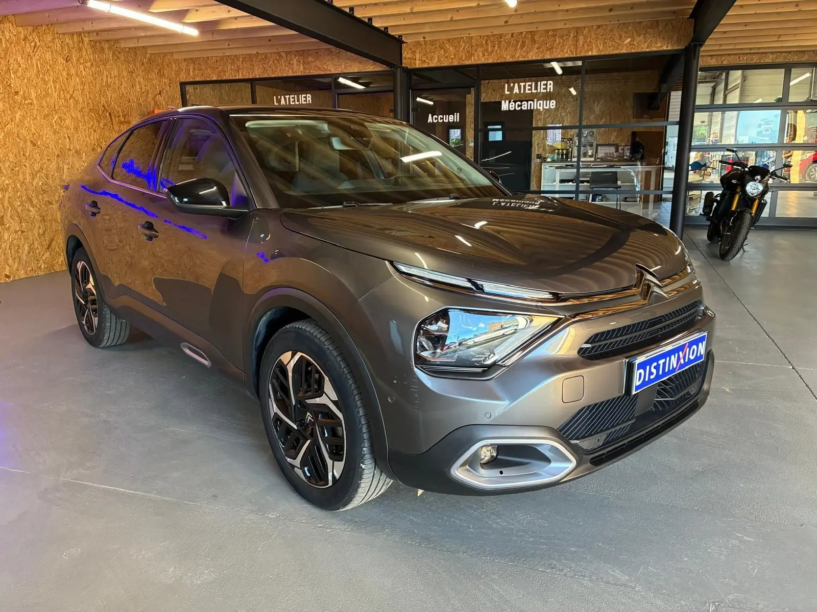 Citroën C4 X gris en 3/4 avant droit dans un atelier, avec jantes noires et éclairage LED visible.