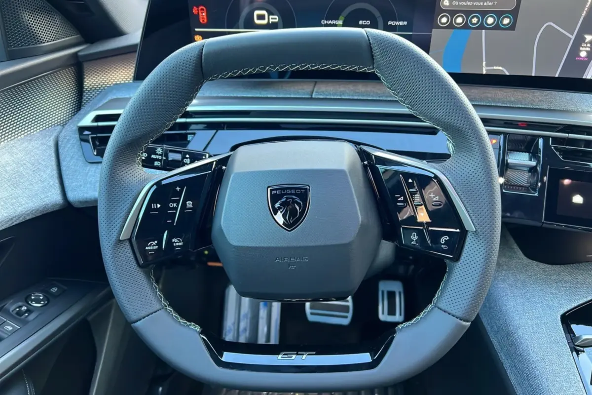 Vue rapprochée du volant cuir gris perforé avec logo Peugeot et inscription GT, tableau de bord numérique en arrière-plan.