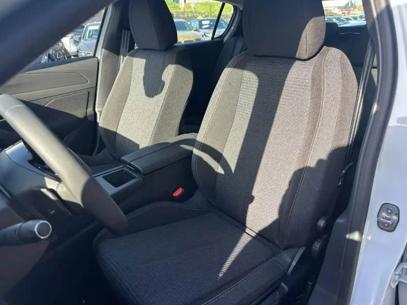Vue intérieure côté conducteur de la Peugeot 308 2025, sièges tissu gris avec accoudoir central et volant multifonction.