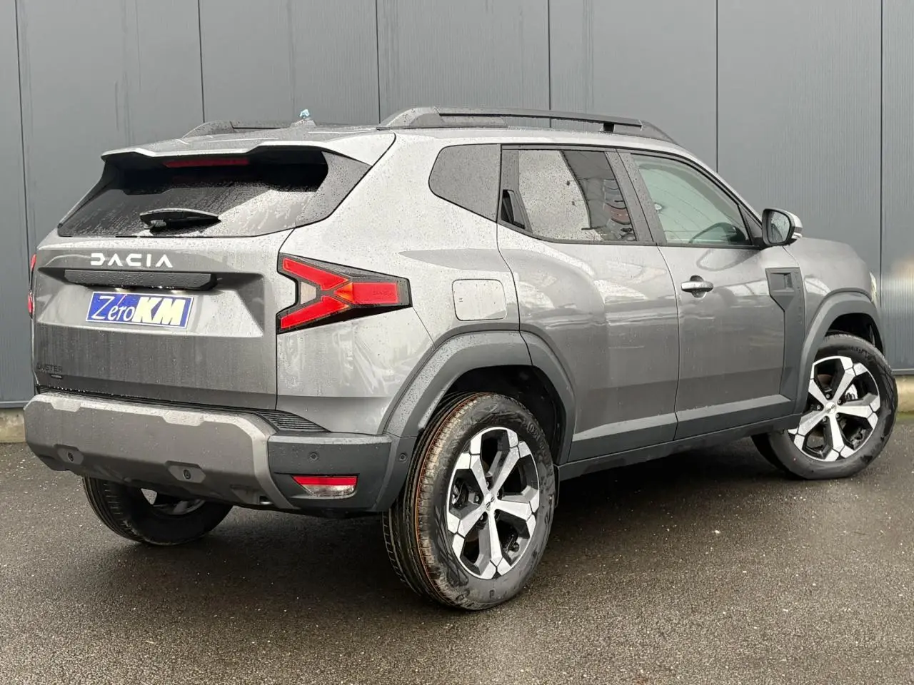 Vue 3/4 arrière droit d'un Dacia Duster gris schiste avec jantes alliage diamantées et barres de toit noires.