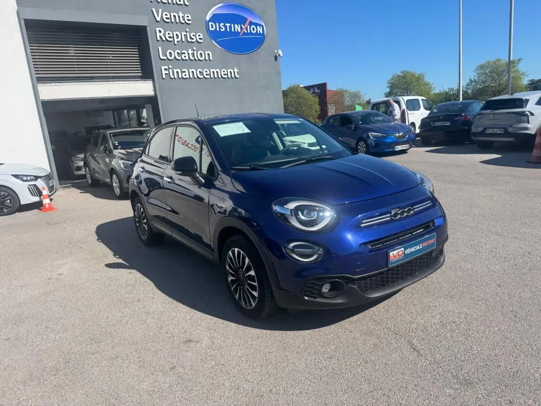 Vue 3/4 avant droit d'une FIAT 500X bleu métallisé avec jantes classiques 16 pouces, stationnée devant un garage.