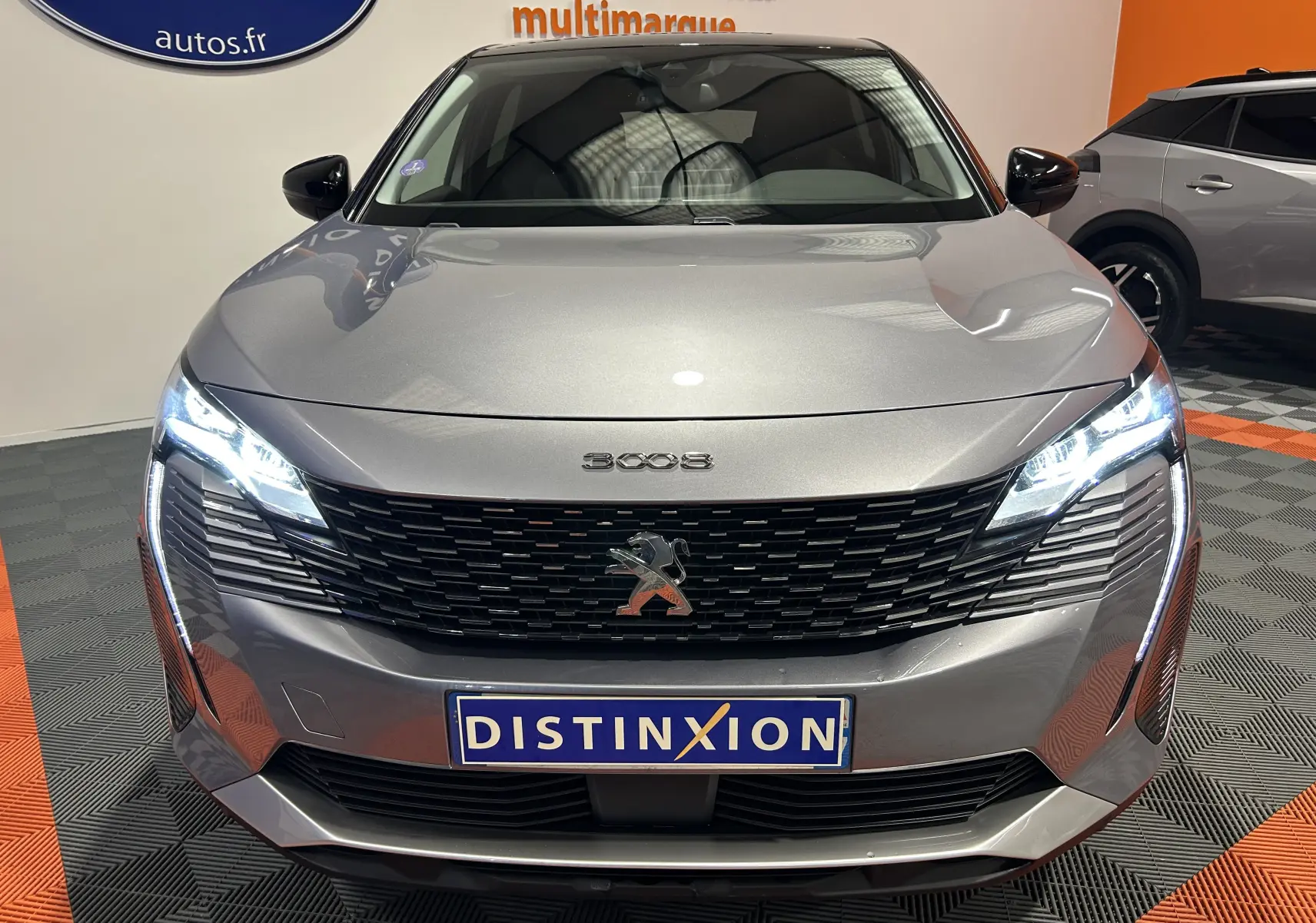 Vue avant d'un Peugeot 3008 gris Artense avec phares allumés et calandre noire distinctive en intérieur showroom.
