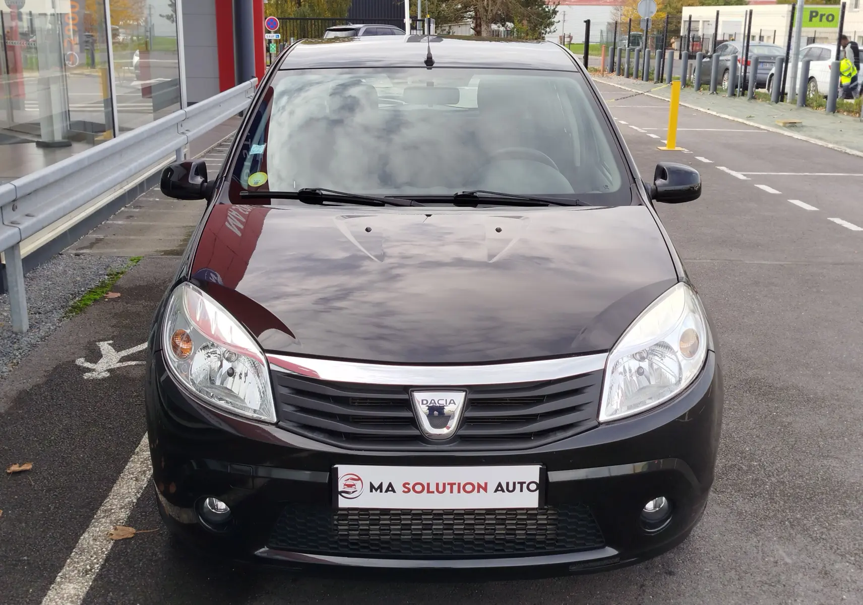 Vue frontale d'une Dacia Sandero noire de 2011 avec calandre chromée et phares allumés sur un parking urbain.