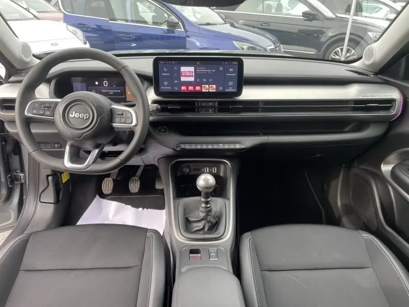Vue intérieure avant du Jeep Avenger 2025 gris métal, montrant le tableau de bord moderne avec écran tactile et boîte manuelle.