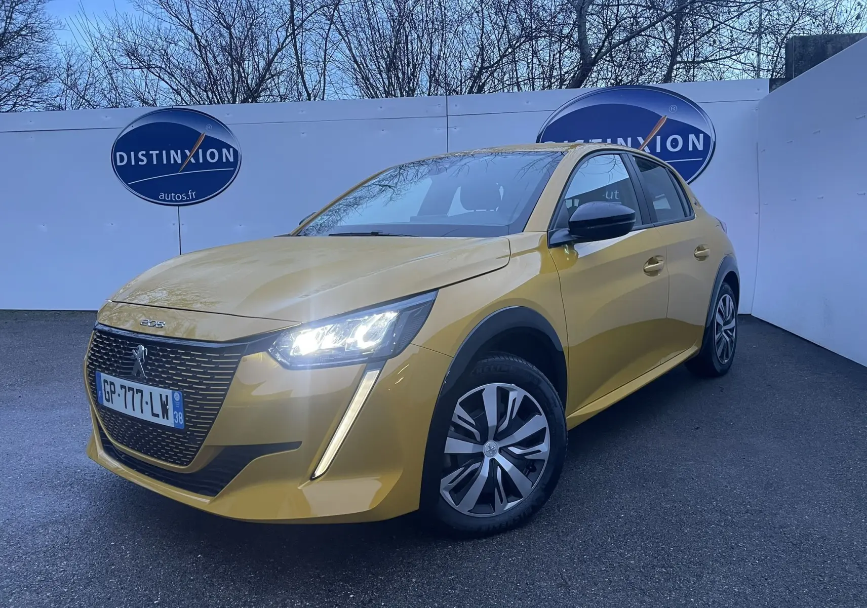 Peugeot 208 électrique jaune vue en 3/4 avant droit avec phares allumés sur fond urbain.