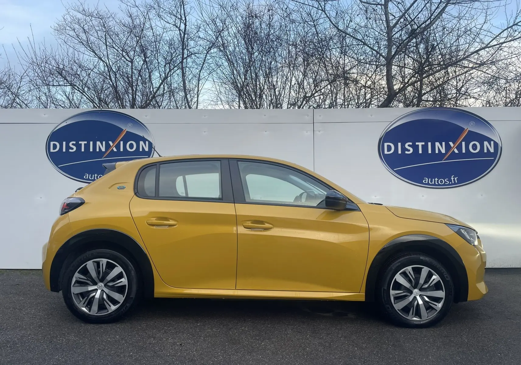 Profil côté gauche d'une Peugeot 208 électrique jaune 2023, avec jantes stylisées et logo Distinxion en arrière-plan.