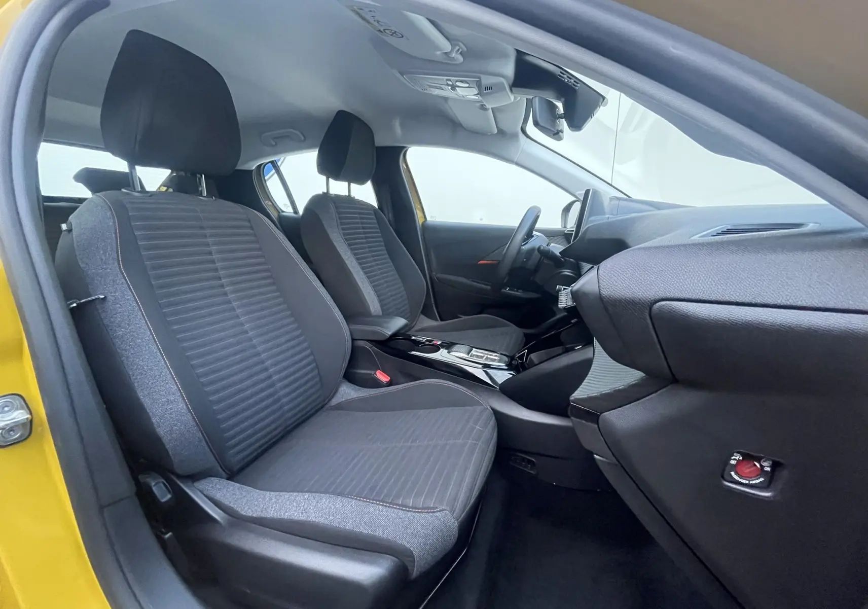Vue intérieure côté conducteur de la Peugeot 208 II électrique jaune 2023, sièges tissu gris et console centrale moderne.