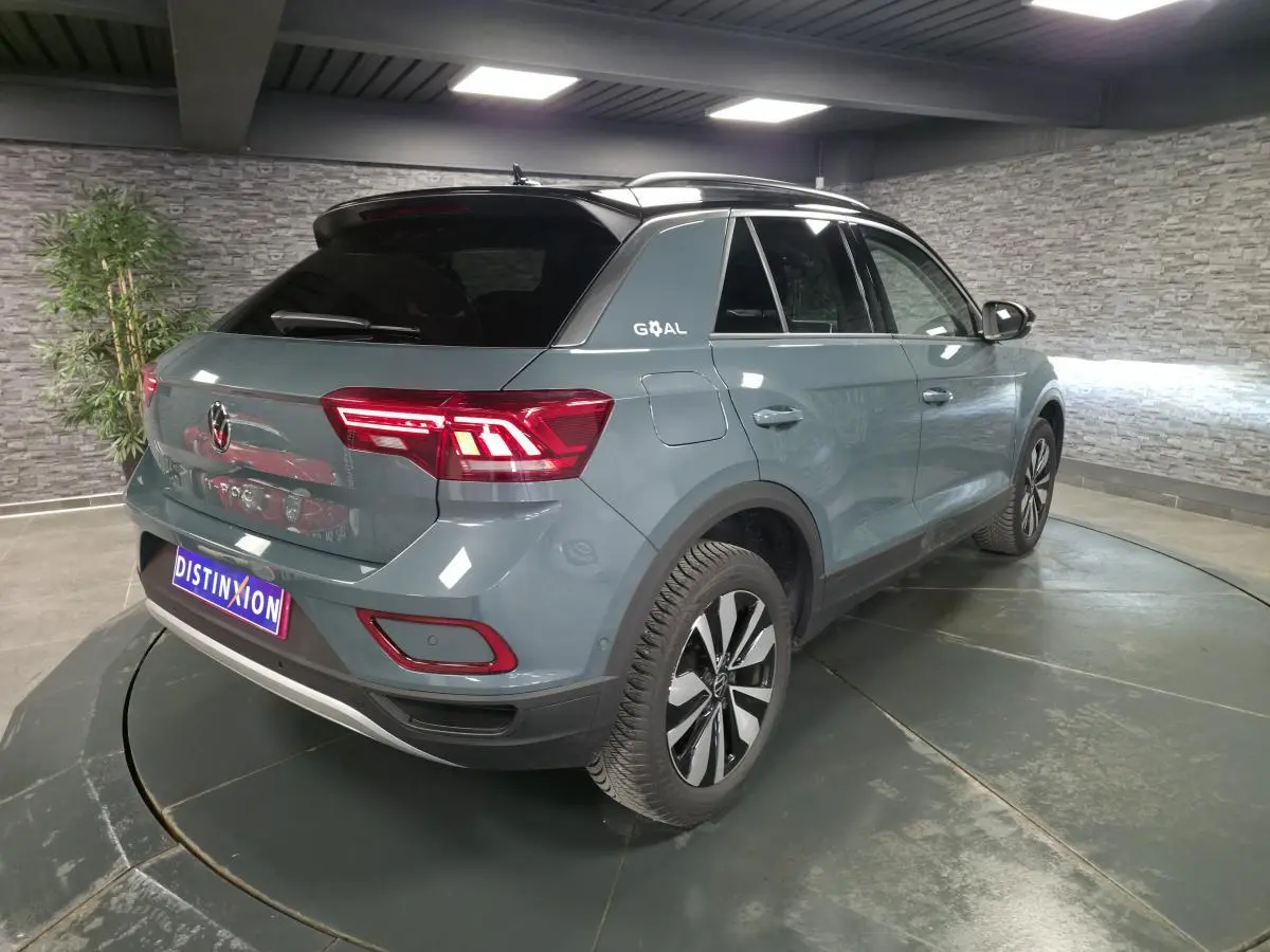 Vue 3/4 arrière droit du Volkswagen T-Roc bleu avec toit noir et feux arrière LED allumés en intérieur.