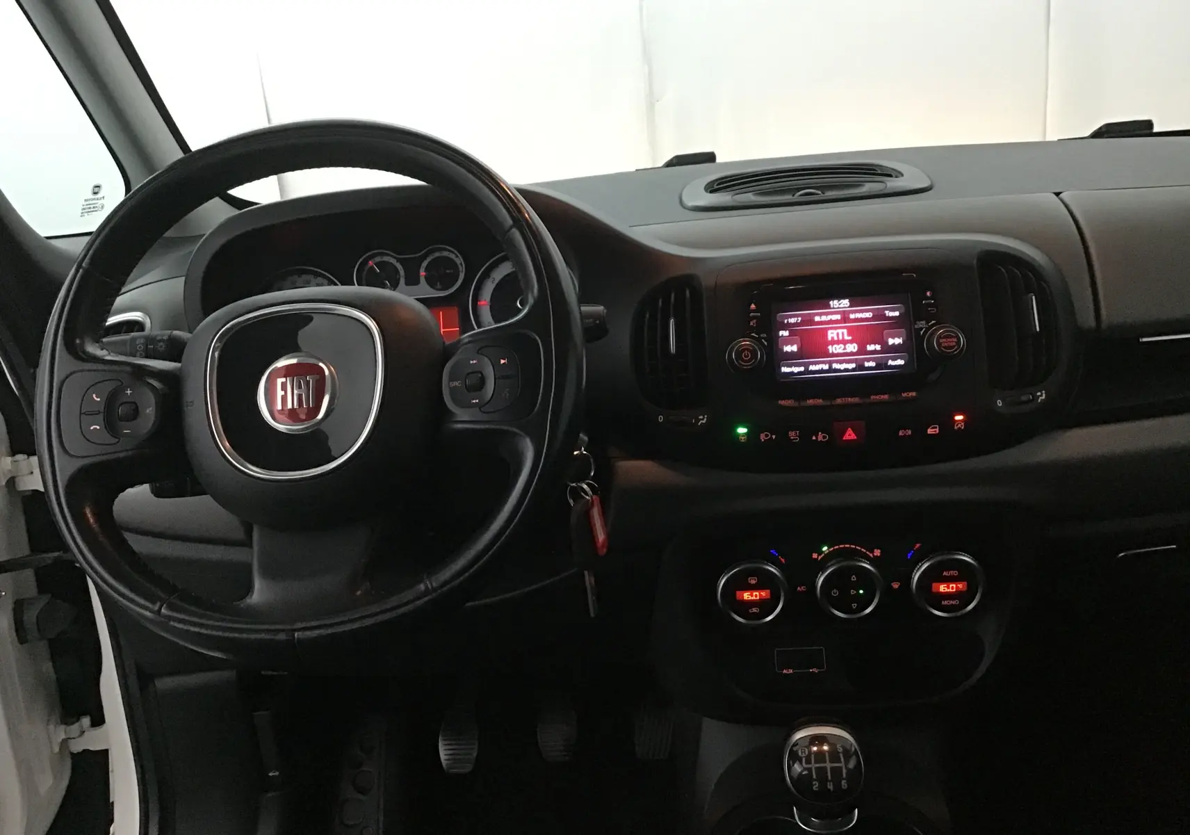 Intérieur de la Fiat 500L blanche vue de face, tableau de bord avec volant, écran multimédia et boîte manuelle visible.