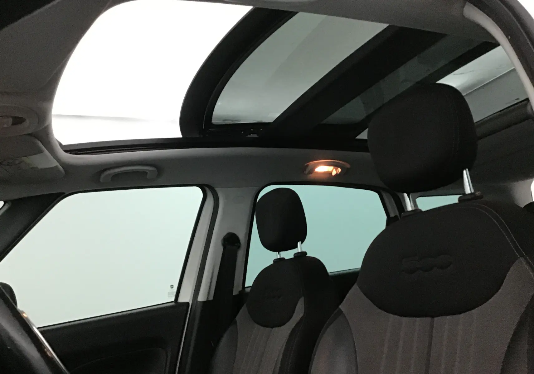 Vue intérieure avant du Fiat 500L blanc 2015, sièges noirs avec logo 500 et toit panoramique ouvert.