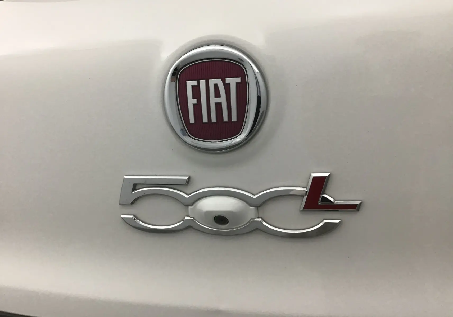 Gros plan sur le logo FIAT et l’inscription 500L chromée avec caméra de recul intégrée sur une carrosserie blanche.