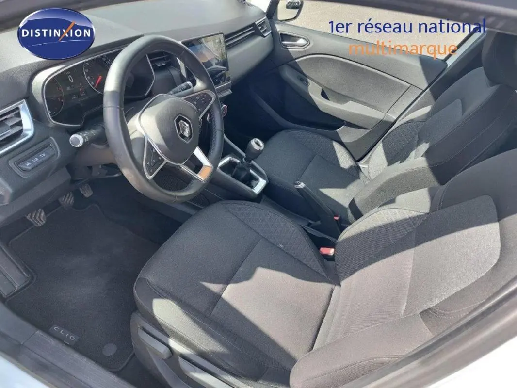 Vue intérieure côté conducteur de la Renault Clio blanche 2021, montrant le volant, la boîte manuelle et les sièges en tissu noir.