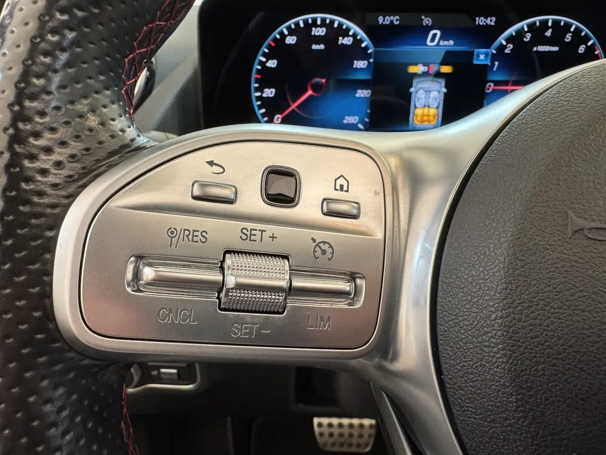 Gros plan sur les commandes argentées du volant cuir perforé noir d’un Mercedes GLA 250 e AMG, tableau de bord numérique visible.